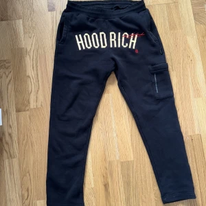 Svarta Hoodrich mjukisbyxor med cargo-ficka - Svarta mjukisbyxor från Hoodrich med stor logga framtill och en praktisk cargo-ficka på ena benet. Byxorna har elastisk midja och är tillverkade i mjukt bomullsmaterial för maximal komfort. Perfekt för en avslappnad streetwear-look.