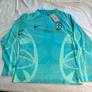 Nike Brasil långärmad fotbollströja L - Snygg långärmad fotbollströja från Nike för Brasilien, i en fräsch ljusblå färg med grafiskt mönster och dragkedja vid halsen. Tröjan har Brasil-logga och Nike Swoosh på bröstet. Tillverkad i lätt och ventilerande funktionsmaterial. Pris kan diskuteras 