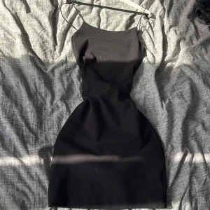 Svart mini dress - En svart, figurnära kort klänning med smala axelband och enkel, stilren design. Klänningen har en rak halsringning och är ärmlös, perfekt för dig som gillar minimalistisk stil och vill ha något klassiskt i garderoben. Aldrig använd endast testad och tvättad 