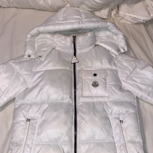 Vit Moncler pufferjacka med huva - Denna snygga jackan kan få vem som helst att se mäktig ut och drar till sig imponerande blickar med dess unika stil. Denna jacka får dig både att sticka ut ur mängden och uppvärmd inför kylan. Den har använts ungefär 5 gånger och tagits bra hand om så den är så gott som ny utan skador. Du har en avtagbar luva och två olika sätt att stänga dragkedjan på, en där Moncler färgerna syns och en där det inte syns. Tveka inte på att höra av dig vid intresse priset kan säkerligen diskuteras. MVH