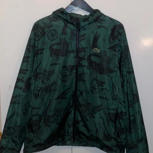 Grön vindjacka Lacoste x Netflix  - Grön vindjacka från Lacoste x Netflix med svart grafiskt mönster av krokodiler och olika symboler. Jackan har huva, hel dragkedja framtill och meshfoder. Perfekt för dig som gillar streetstyle och unika samarbeten.