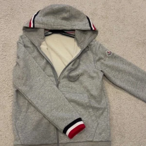 Grå Moncler hoodie med dragkedja - Snygg grå hoodie från Moncler med hel dragkedja, huva och ribbade muddar i rött, vitt och marinblått. Tröjan har Moncler-logga på ärmen och praktiska fickor framtill. Perfekt för dig som gillar stilrena och sportiga plagg. I helt nyskick påse tillkommer även, pris kan diskuteras vid snabb affär. Tveka inte på höra av dig.