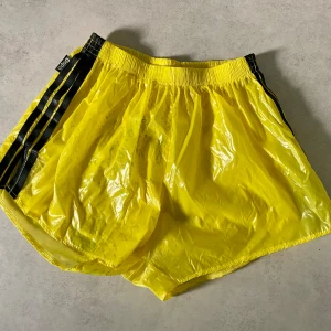 Gula shorts från Dapa Designs, Large - Säljer ett par riktigt coola gula shorts från Dapa Designs i storlek Large. De har en glansig finish, svarta ränder på sidorna och elastisk midja. Materialet är syntet och de har en loose passform, perfekt för träning eller chill dagar. Några missfärgningar förekommer, se bilder 