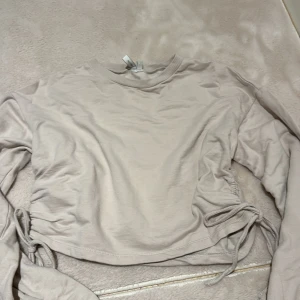 Beige långärmad croppad topp med snörning - Säljer en beige långärmad croppad topp från Gina Tricot. Toppen har rund halsringning, ribbade muddar vid ärmsluten och snygga snörningsdetaljer i sidorna som ger en trendig look. Perfekt att styla med höga jeans eller kjol.