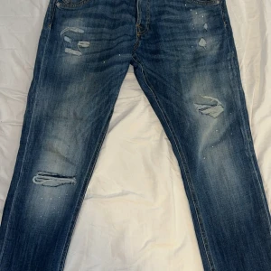 Jack & Jones jeans - Snygga blå jeans med slitningar och hål på benen samt coola färgstänk från Jack & Jones. Jeansen har raka ben och klassisk femficksdesign. Perfekta för en avslappnad streetstil.