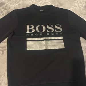 Svart tröja med silvertryck från Hugo Boss - Svart långärmad tröja från Hugo Boss med stort silverfärgat BOSS-tryck och bred silverdetalj på bröstet. Tröjan har en klassisk rund halsringning och är tillverkad i ett mjukt material som känns skönt mot huden. Perfekt för dig som gillar stilrena och moderna plagg.