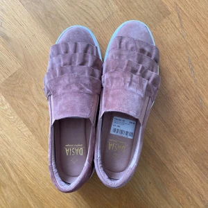 Rosa mockasneakers från Dasia - Säljer ett par snygga sneakers från Dasia i rosa mocka. Skorna har en platt vit sula och dekorativa volanger framtill som ger en unik look. Perfekta för dig som vill ha något extra till vardagsstilen.