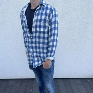 Blåvit rutig skjorta i bomull - Säljer en blåvit rutig skjorta i mjuk bomull. Skjortan har klassisk krage, knäppning framtill och en bröstficka. Långärmad modell med avslappnad passform, perfekt att bära öppen över en t-shirt.