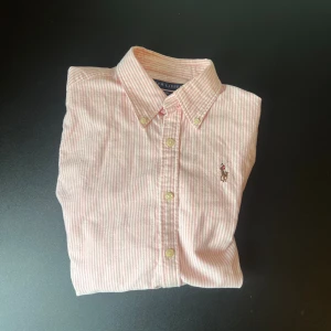Rosa randig skjorta från Ralph Lauren - Säljer en slim fit skjorta från Ralph Lauren i rosa och vitt med tunna ränder. Skjortan har klassisk krage, knäppning framtill och den ikoniska broderade loggan på bröstet. Perfekt för dig som gillar preppy stil och snygga detaljer.
