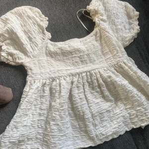 Vit blus med puffärmar från Gina Tricot - Supersöt vit blus från Gina Tricot med puffiga korta ärmar och strukturerat tyg. Blusen har en lös passform och en lätt rynkad nederdel som ger en flowy känsla. Perfekt att styla med jeans eller kjol för en trendig look.