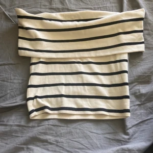 Randig offshoulder tubtopp - Snygg offshoulder tubtopp i beige med svarta horisontella ränder. Toppen har en bred kant upptill som ger en cool och avslappnad look. Perfekt att matcha med jeans eller kjol för en trendig stil.