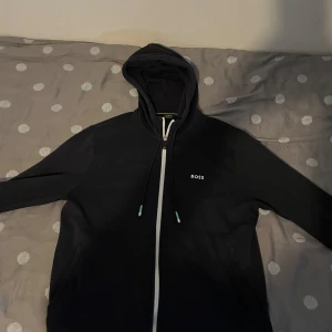 Svart hoodie från BOSS med dragkedja - Svart hoodie från BOSS med vit dragkedja och snörning i huvan. Liten BOSS-logga på bröstet och upprepade BOSS-tryck längs nederkanten på baksidan. Perfekt för en chill och stilren look.