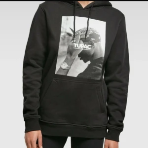 Svart Tupac hoodie med tryck - Svart hoodie med stort Tupac-tryck på bröstet. Tröjan har huva med snörning och en klassisk magficka. Perfekt för dig som gillar streetstyle och vill ha en chill look. Materialet känns mjukt och bekvämt mot huden. Passar både till killar och tjejer.