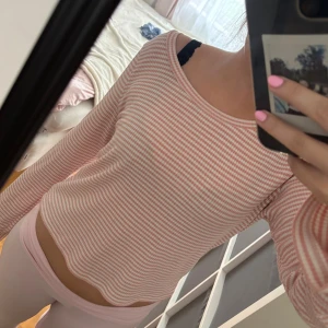 Randig rosa och vit långärmad tröja - Säljer en söt långärmad tröja med smala rosa och vita ränder. Tröjan har en rund halsringning och är lite croppad med vågig nederkant. Perfekt för dig som gillar en chill och feminin stil.