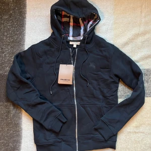 Svart hoodie från Burberry med dragkedja - Svart hoodie från Burberry med klassiskt rutigt foder i huvan, broderad logga på bröstet och hel dragkedja framtill. Tröjan har snörning i huvan och två fickor. Tillverkad i mjuk bomull för en skön känsla.