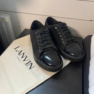 Svarta sneakers från Lanvin i mocka(byten funkar) - Snygga svarta sneakers från Lanvin med ovandel i mocka och lackad tå. Skorna har rund tå, snörning och platt sula. Klassisk och stilren design med exklusiv känsla, perfekt för dig som gillar lyxiga detaljer. Kommer med originalpåse från Lanvin. Og påse medföljer vid köp. Billigare vid snabba affär. Hör av er vid minsta lilla. Mochan är rätt sliten 
