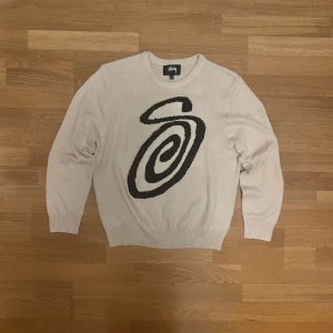 Beige stickad tröja från Stussy - Vit stickad tröja från Stussy med ett stort svart grafiskt mönster på framsidan. Tröjan har rund halsringning, ribbade muddar och är långärmad. Perfekt för dig som gillar streetwear och vill sticka ut med en unik design.