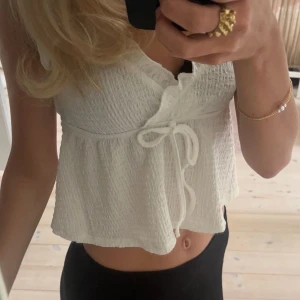 Vit croppad blus med knyt - Supersöt vit croppad blus med knyt framtill och lätt rynkad struktur. Toppen har v-ringning och volangkant vid halsen, vilket ger en romantisk vibe. Perfekt till jeans eller kjol för en fräsch look.
