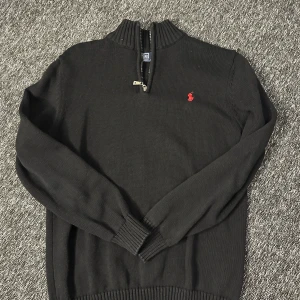 Svart stickad tröja Polo Ralph Lauren - Svart stickad tröja från Polo Ralph Lauren med halv dragkedja och röd broderad logga på bröstet. Tröjan har ribbad krage, muddar och nederkant samt lång ärm. Perfekt för lager-på-lager och snygg till jeans eller chinos.
