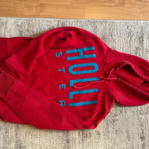 Röd hoodie från Hollister med blå text - Röd hoodie från Hollister med stor blå broderad logga framtill. Klassisk känguruficka, dragsko i huvan och mjukt bomullsmaterial. Perfekt för dig som gillar en chill och sportig stil. Långärmad och normal passform.