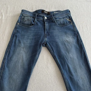 Blå Replay Anbass jeans, midwaist - Snygga blå Replay Anbass jeans med klassisk femficksdesign och diskreta slitningar. Jeansen har en rak passform och är tillverkade i mjukt denimtyg. Märkesdetaljer syns på bakfickan och patchen i midjan. Perfekta för en avslappnad och trendig look. Liten, liten fläck syns på sista bilden.