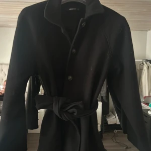 Svart kappa med bälte från Gina Tricot - Stilren svart kappa från Gina Tricot med knappar framtill och knytskärp i midjan. Klassisk krage och långärmad modell som passar perfekt till höst och vinter. Enkel och tidlös design som är lätt att matcha.