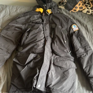 Svart Parajumpers dunjacka med huva - Svart dunjacka från Parajumpers med huva, flera fickor och ikonisk gul rem vid kragen. Jackan har patch på ärmen och dragkedjor framtill. Perfekt för kalla dagar och har en sportig, urban vibe.