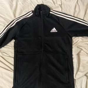 Svart Adidas track jacket med vita ränder - Svart Adidas track jacket med klassiska vita ränder längs ärmarna och dragkedja framtill. Jackan har hög krage, två fickor och Adidas-logga på bröstet. Tillverkad i polyester och har AEROREADY-tryck på ryggen. Perfekt för sportiga vibes.