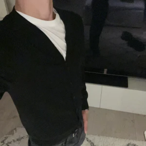 Svart kofta från J.Lindeberg - Stilren svart kofta från J.Lindeberg med v-ringning och knappar framtill. Långärmad modell i mjuk bomull, perfekt att slänga över en t-shirt för en clean look. Passar dig som gillar klassisk och enkel stil. Säljer för att den är för liten och pris går såklart att diskutera!