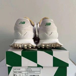 New Balance x Casablanca XC-72 sneakers - Säljer ett par New Balance x Casablanca XC-72 sneakers i vitt och ljusgrått med gröna och orange detaljer. Skorna har en chunky, mönstrad sula, perforerade paneler och snörning med guldfärgade detaljer. Materialmix av mocka och syntet för en sportig och trendig look.