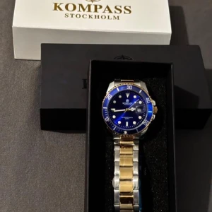 Blå och guld herrklocka Kompass - Snygg herrklocka från Kompass Stockholm med blå urtavla och vridbar bezel. Armbandet är i rostfritt stål med guld- och silverfärgade länkar. Klockan har datumvisning och levereras i originalask med garanti.