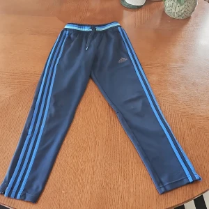 Adidas blå träningsbyxor barn 140 - Mörkblå träningsbyxor från Adidas med ljusblå ränder längs benen och snörning i midjan. Klassisk Adidas-logga på låret och elastisk midja. Perfekta för sport eller chill. Tillverkade i polyester för skön passform.