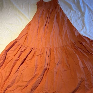 Lång orange sommarklänning - Snygg långklänning i en härlig orange färg med smock upptill och breda axelband. Klänningen har en luftig och flowy passform med volangdetalj nedtill, perfekt för varma dagar.