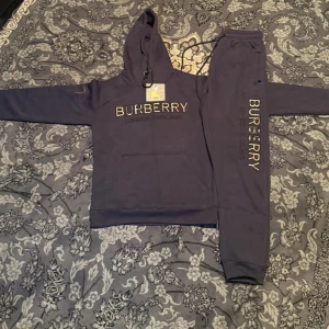 Mörkblå Burberry hoodie med brodyr - Säljer en mörkblå hoodie från Burberry med stor broderad logga i beige, rött och svart på bröstet. Tröjan har huva med snörning och en klassisk känguruficka framtill. Mjuk bomullskänsla och långärmad modell, perfekt för höst och vinter.