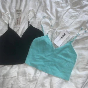 Ribbad topp med pärlband  - Säljer två ribbade crop tops från FB Sister i storlek M (passar S)och S. En är svart och en är turkos, båda med unika axelband av pärlor och små fjärilar. Topparna har v-ringning och är ärmlösa, perfekta för att sticka ut med en cool detalj på axelbanden.