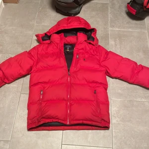 Röd pufferjacka från Polo Ralph Lauren XL - Riktigt snygg röd pufferjacka från Polo Ralph Lauren i storlek XL i barn passar S. Jackan har huva, två dragkedjefickor och klassisk Polo-logga på bröstet. Perfekt för kalla dagar med sin vadderade design och sportiga look. Stängs med dragkedja framtill. Pris kan diskutera 
