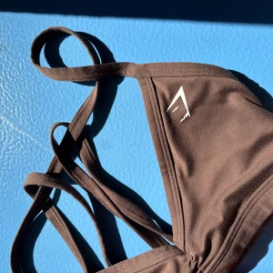 Brun gym topp  - Snygg brun gym överdel från gymshark med trekantsformade kupor och flera tunna axelband för en trendig look. Mjuk och stretchig i materialet, perfekt för gymmet . Vit logga framtill ger en sportig touch. Detta är storlek xs. Justerbara band i nacken!  Denna förgen finns inte längre  att köpa på gymshark! 