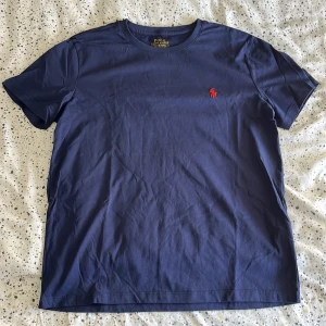 Marinblå t-shirt Polo Ralph Lauren - Snygg marinblå t-shirt från Polo Ralph Lauren med röd broderad logga på bröstet. Modellen är slim fit och har klassisk rund hals och korta ärmar. Tillverkad i mjuk bomull som känns skön mot huden.