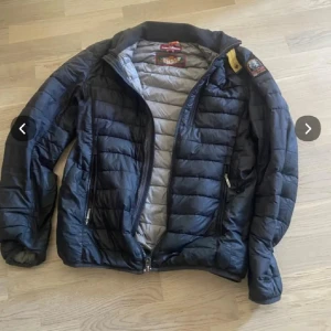 Svart dunjacka från Parajumpers - Snygg svart dunjacka från Parajumpers med klassisk patch på ärmen och gul detalj vid axeln. Jackan har quiltad design, två dragkedjefickor framtill och hög krage. Perfekt för kalla dagar och riktigt skön att bära. Dragkedja saknar en grej som man drar upp på den övre delen men går fof att dra upp inte jättebra men funkar ändå och den är ganska sliten med små hål och trådar