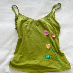 Grön linnetopp med broderade blommor - Säljer en grön linnetopp med smala axelband och v-ringning. Toppen har broderade blommor i rosa, gul och blå längs ena sidan.