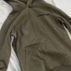 Olivgrön hoodie från Calvin Klein Jeans - Olivgrön hoodie från Calvin Klein Jeans med svart logga på bröstet och en snygg logoremsa längs ryggen. Tröjan har känguruficka, huva och är tillverkad i en mjuk bomullsblandning. Storlek är 16 (ålder)  Skriv vid frågor😀
