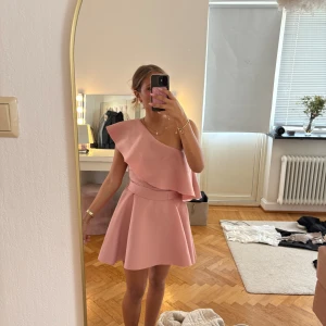 Rosa one shoulder volangklänning - Supersöt ljusrosa kortklänning med one shoulder och stor volangdetalj över axeln. Klänningen har en markerad midja och en lätt utsvängd kjol. Snygg rosettdetalj baktill. Perfekt för dig som vill sticka ut med en feminin och trendig look.