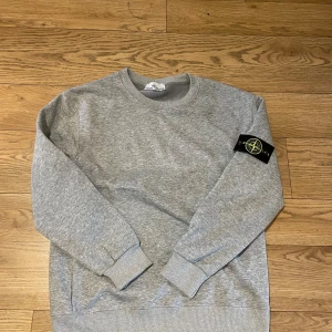 Grå sweatshirt från Stone Island - Grå crewneck sweatshirt från Stone Island med klassisk logga på vänster ärm. Tröjan har ribbade muddar vid ärmslut och nederkant samt rund halsringning. Tillverkad i mjuk bomull för en skön och avslappnad stil.