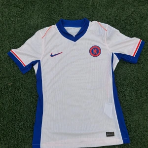 Chelsea FC vit bortatröja Nike M - Chelsea FC:s officiella bortatröja från Nike i vitt med blå och röda detaljer. Tröjan har korta ärmar, blå V-ringad krage och ventilerande Dri-FIT ADV-material. Broderat klubbmärke på bröstet och diskret strukturmönster i tyget. Perfekt för dig som älskar fotboll och Chelsea.