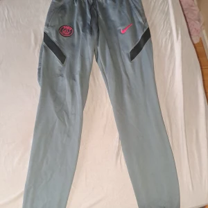 Grå PSG träningsbyxor från Nike - Grå träningsbyxor från Nike med PSG-logga och rosa swoosh på framsidan. Byxorna har svarta detaljer vid fickorna och är tillverkade i Dri-FIT-material med 91% polyester och 9% elastan. Perfekta för fotboll eller träning.