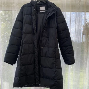Svart lång pufferjacka från 157 - Svart lång pufferjacka från 157 med huva och quiltad design. Jackan har dragkedja och tryckknappar framtill, perfekt för kalla dagar. Klassisk och stilren look som funkar till det mesta. Materialet är syntet och jackan är vadderad för extra värme. Passar XS/S nypris ligger på 500
