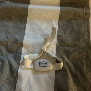 Hov1 armband - Snyggt vitt armband med ett stilrent kronmotiv på en rektangulär platta. Armbandet har ett glansigt band som knyts och ger en cool och enkel look. Perfekt accessoar för dig som gillar minimalistiska detaljer. 