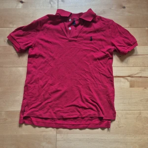 Röd pikétröja från Polo Ralph Lauren - Klassisk röd pikétröja från Polo Ralph Lauren med korta ärmar, krage och två knappar framtill. Tröjan har den ikoniska broderade loggan i mörkblått på bröstet och är tillverkad i bomull för en skön och luftig känsla. Kontakta gärna vid frågor!