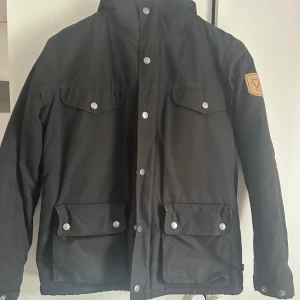 Fjällräven Greenland Jacka - Svart jacka från Fjällräven i storlek XS med mysigt teddyfoder. Klassisk design med fyra fickor framtill, tryckknappar och dragkedja. Snyggt märke på ärmen och praktisk huva. Perfekt för kyliga dagar och outdoor vibes.