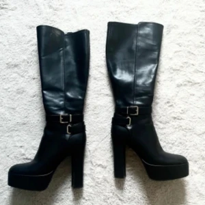 Svarta höga stövlar med klack - Snygga svarta stövlar i skinnimitation med hög klack och platåsula. Stövlarna har rund tå och dekorativa spännen i silver vid ankeln. Perfekta för att lyfta vilken outfit som helst med en edgy vibe och är 13cm högklack.